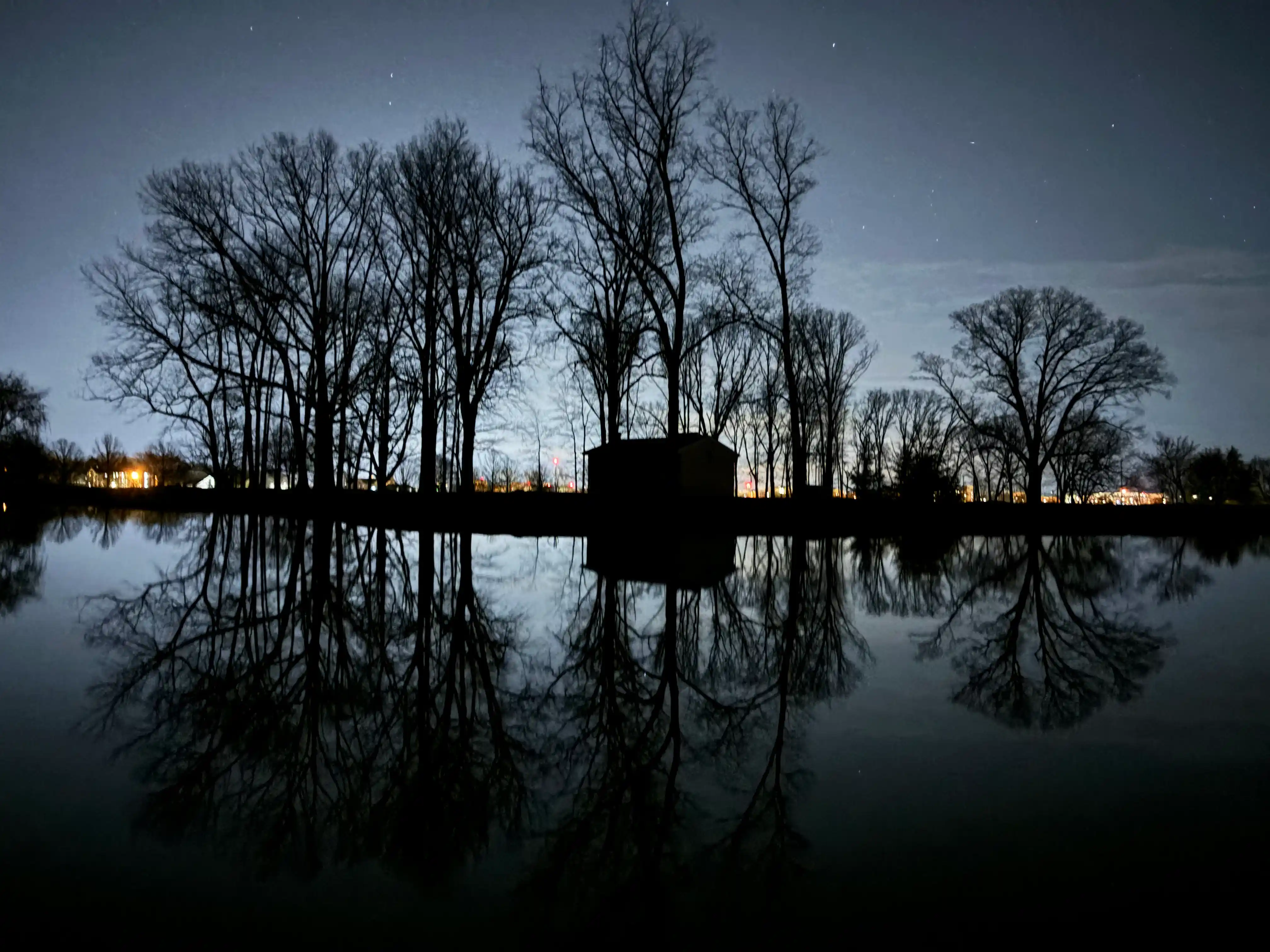 A pond, seen at night, covers the entire foreground of the image; in the far background, there is some city lighting. Immediately behind the pond, there is a collection of trees and a small building, the reflections of which can be seen in the water of the pond.