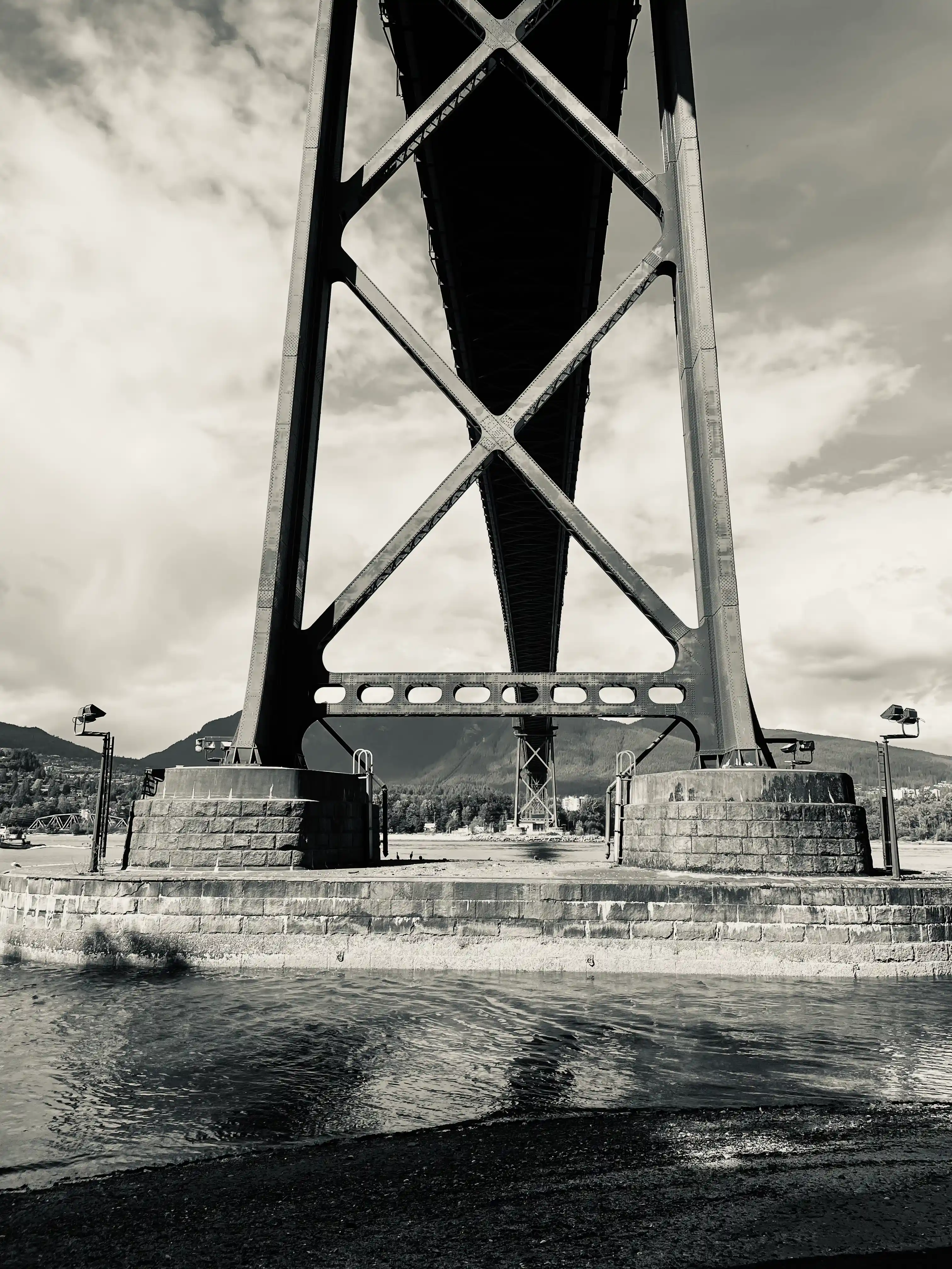 Lions Gate Bridge is seen from below in Stanley Park. In the foreground, there is some gravel before the beginning of the water of Vancouver Harbor. The center of the image contains a stone base supporting two columns of steel with tresses between them. Behind these columns, the bridge stretches overhead.