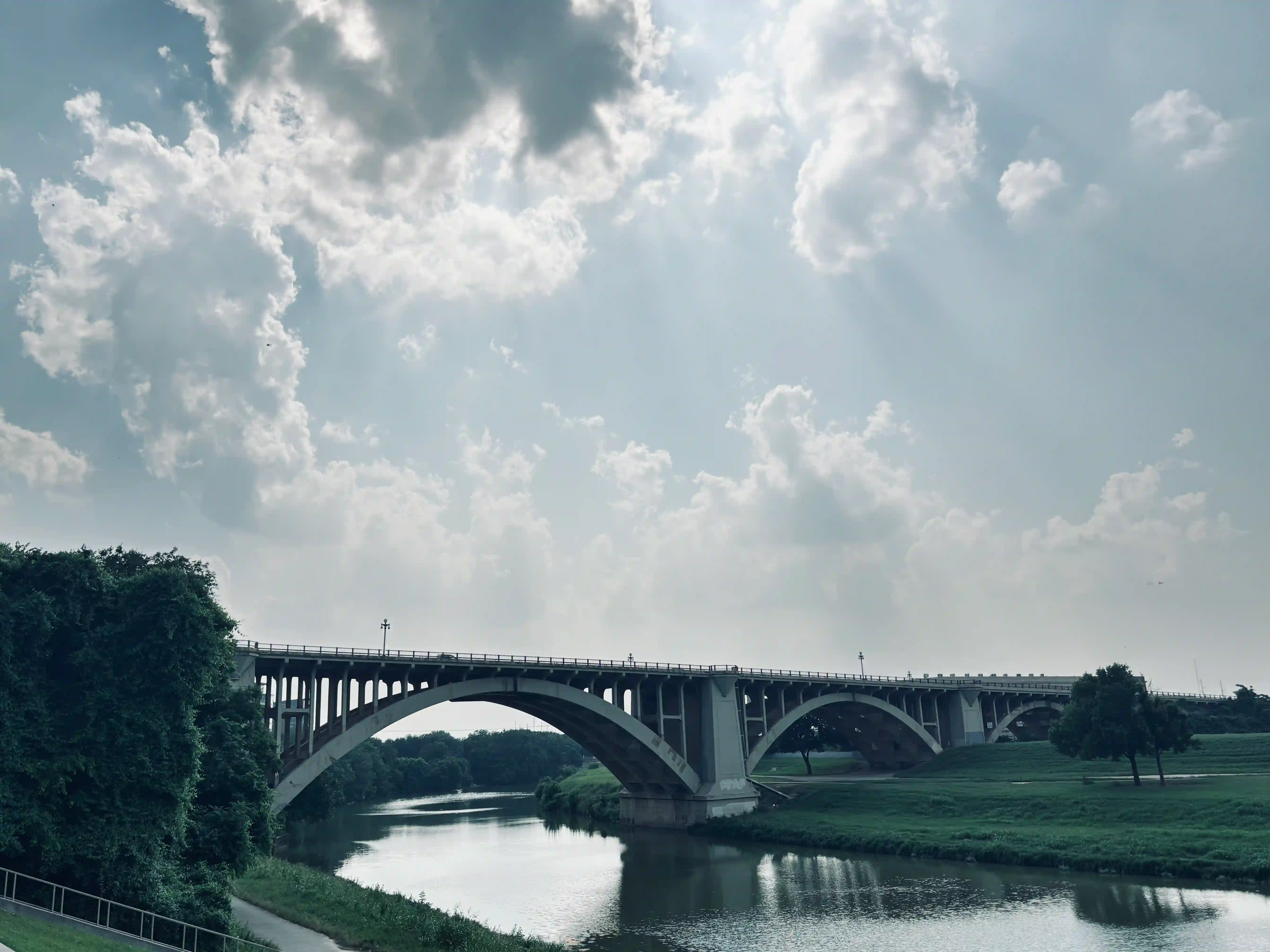 A bridge across the West Fork Trinity River near downtown Fort Worth, Texas. Above, there is a cloudy sky with streaks of sunlight coming through.