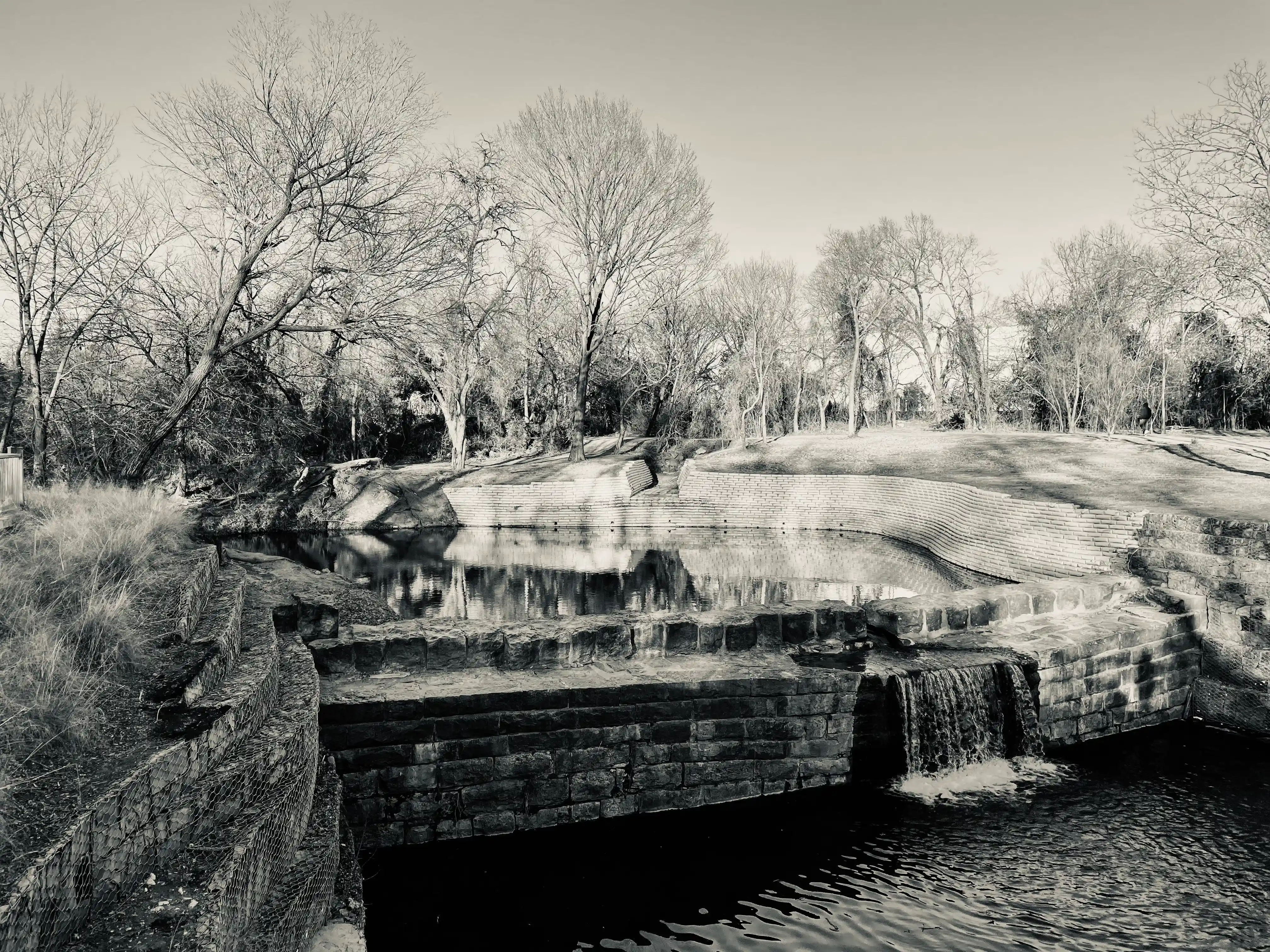 A black and white image of the historic Old Stone Dam of Allen is seen; the dam is in the center of the image, with the waters of Cottonwood Creek flowing through the dam occupying much of the central third of the image. Some shrubs are visible in the foreground to the left, and there are several leafless trees behind the creek. 