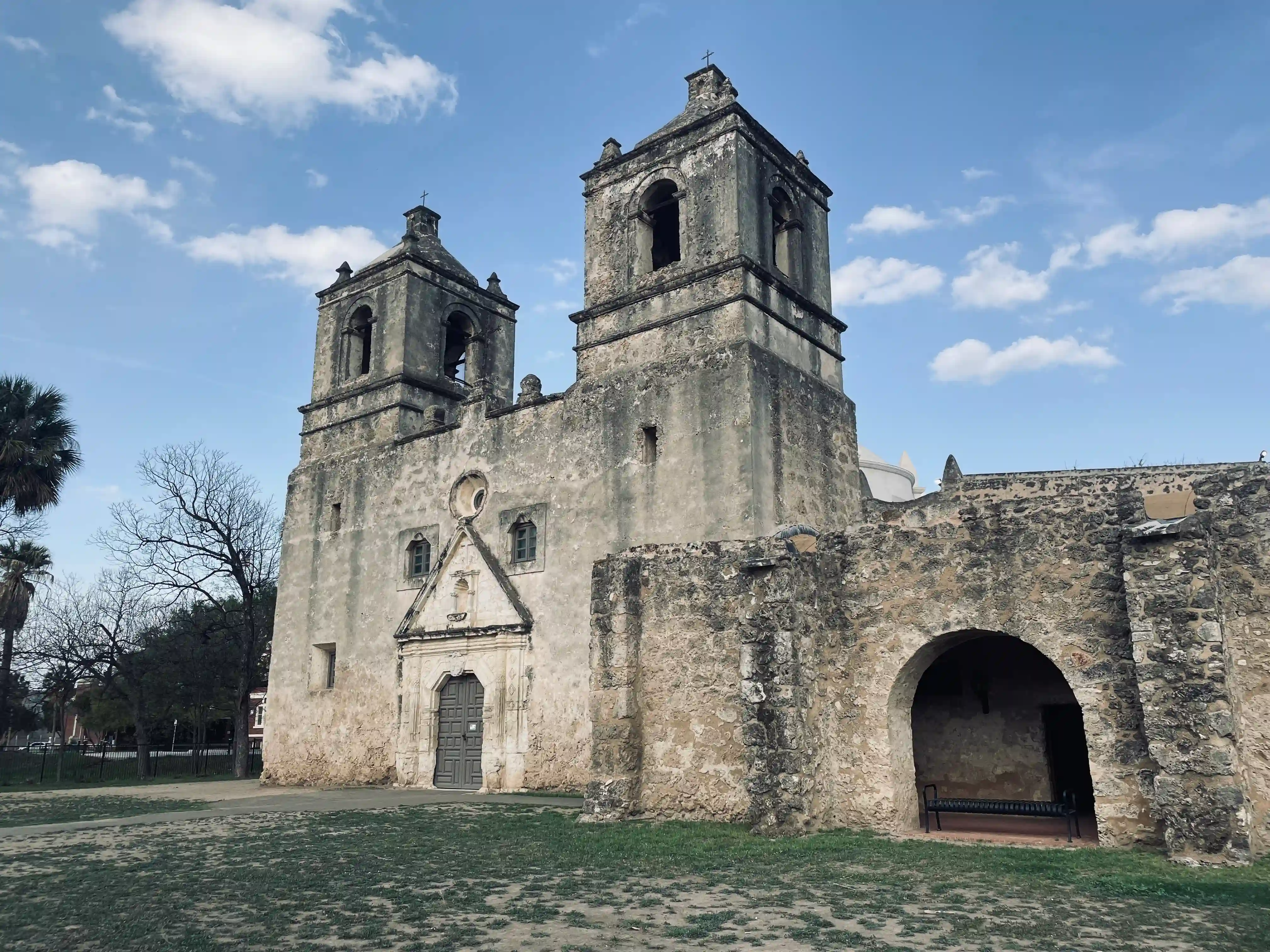 The Church at Mission Nuestra Señora de la Purísima Concepción de Acuña. At the foreground of the image, there are some grasses and dirt, behind which the old church building, constructed of stone, stands. In the image, two towers of the church building are visible to the center. Below the towers, a door into the church can be seen. Towards the right, an arch is visible.