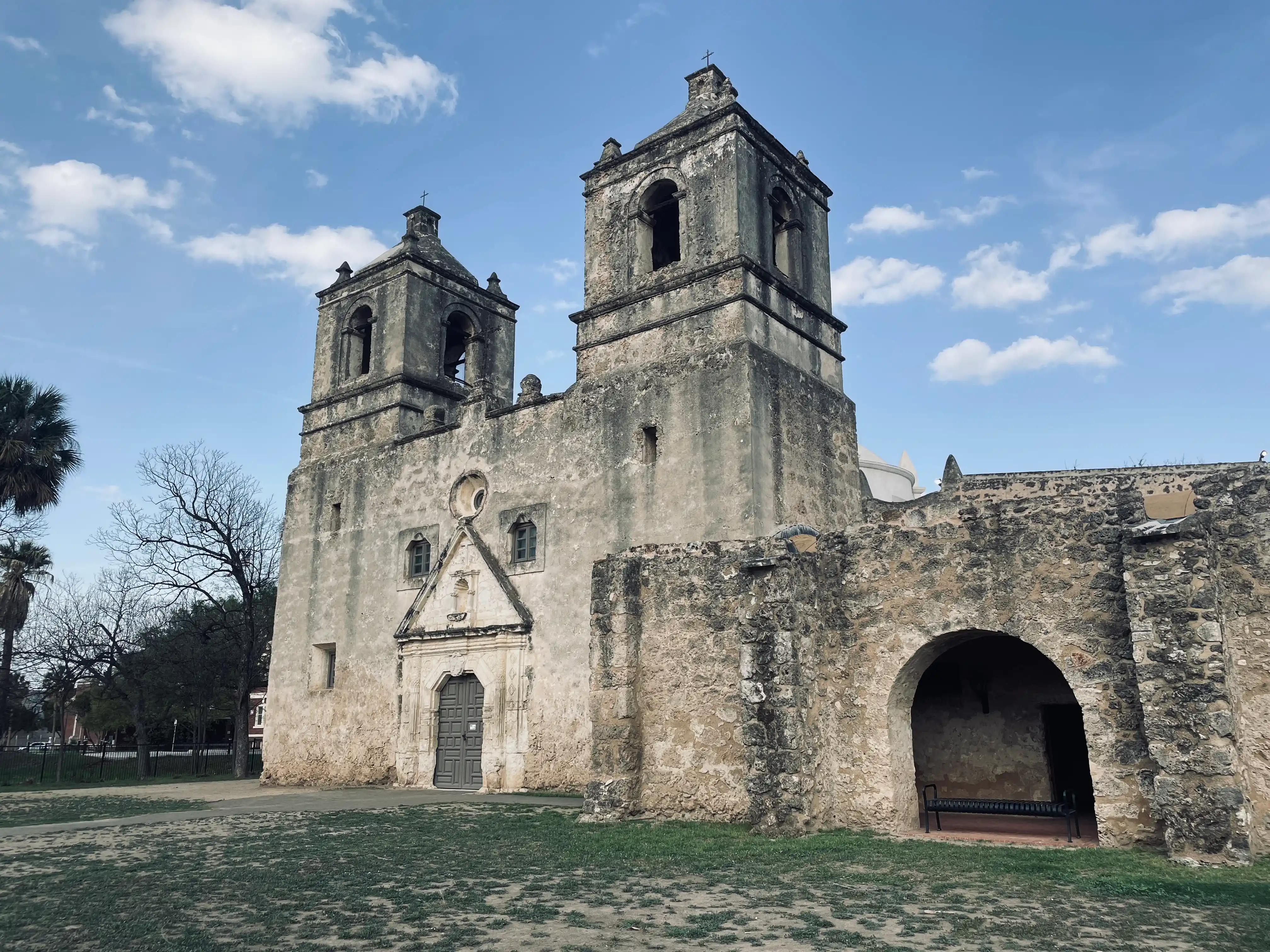 The Church at Mission Nuestra Señora de la Purísima Concepción de Acuña. At the foreground of the image, there are some grasses and dirt, behind which the old church building, constructed of stone, stands. In the image, two towers of the church building are visible to the center. Below the towers, a door into the church can be seen. Towards the right, an arch is visible.