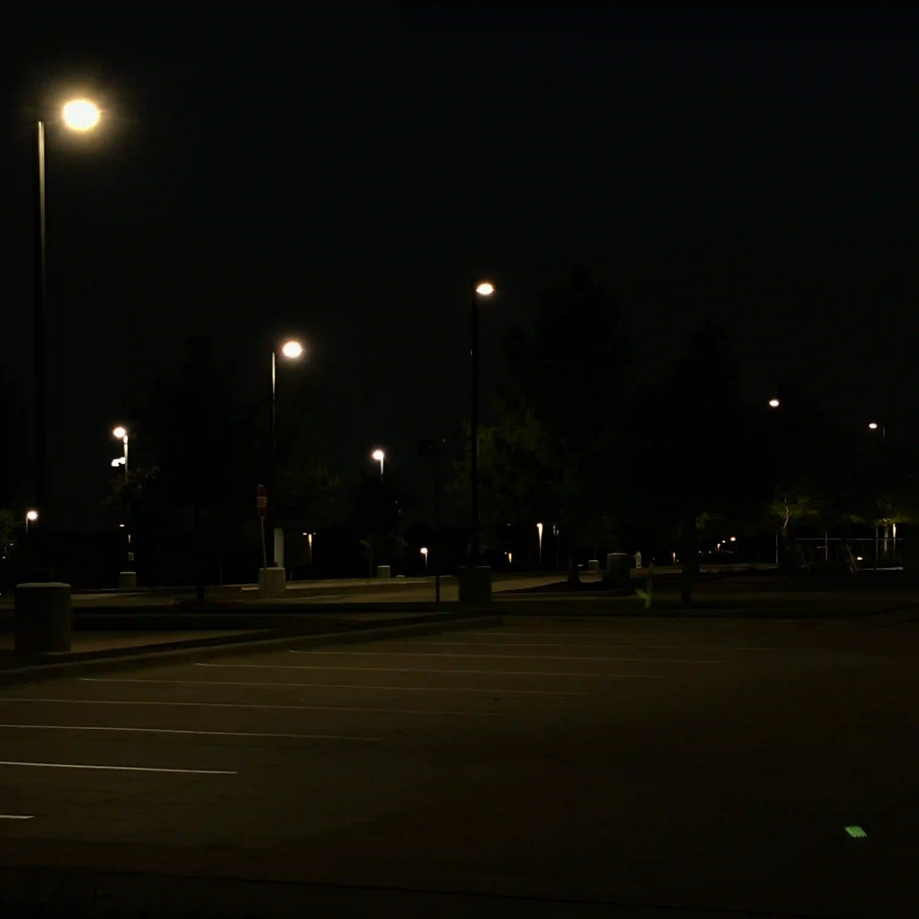 A parking lot at night; dim streetlights illuminate the empty lot in a somewhat eerie manner.