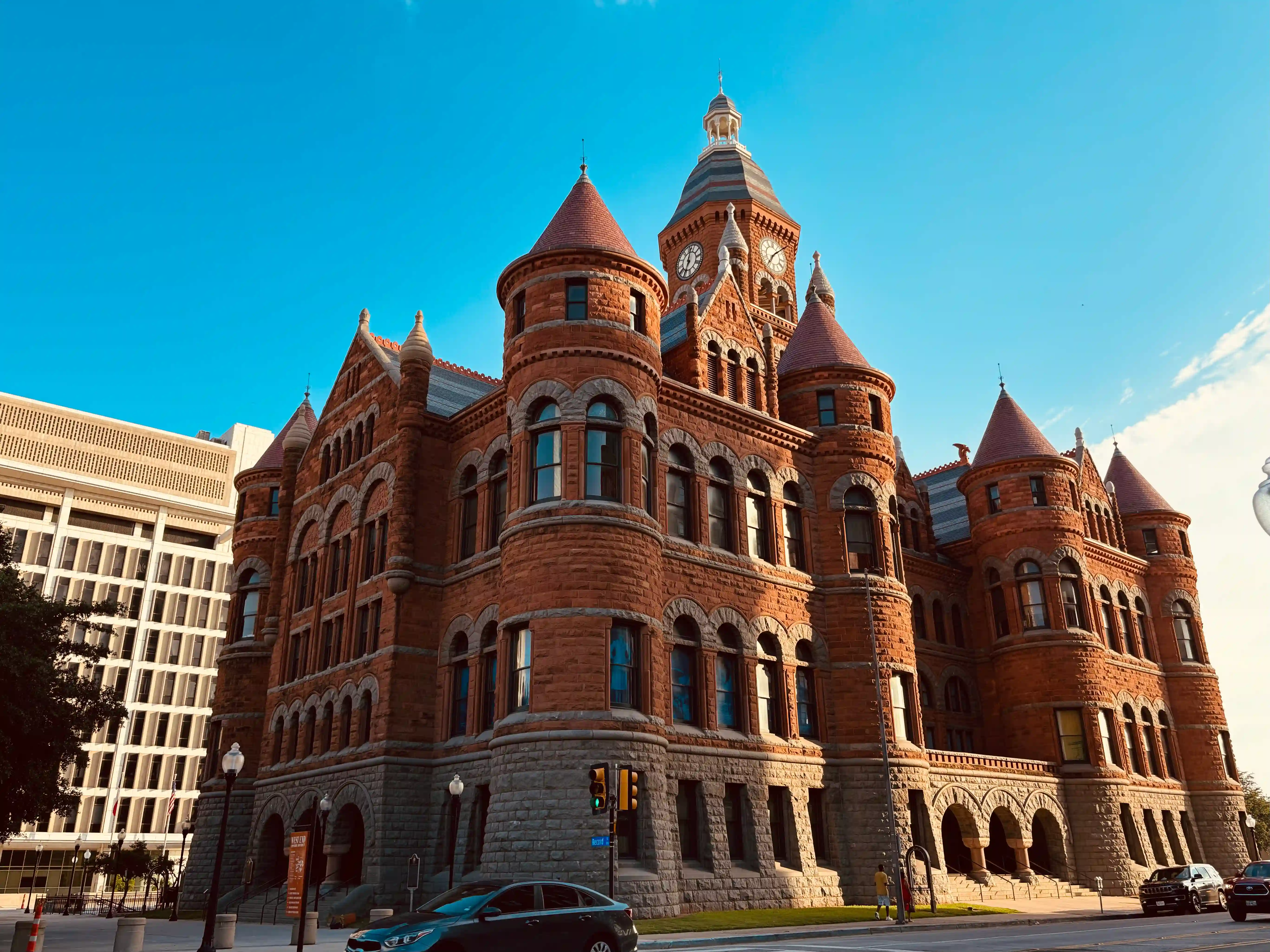 The Old Red Courthouse of Dallas is seen in the center of the picture. In the foreground, there are a few cars, and in the background the Dallas County Records office is visible.